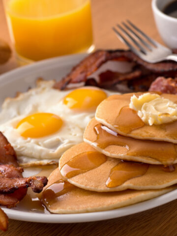 white plate with breakfast pancakes, bacon, eggs, coffee and more.